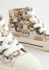 Zapatillas altas beige con estampado de osos, cordones blancos, cremallera lateral y suelas de goma. Material de lienzo texturizado y detalles de costura en contraste.