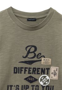 Oliwkowa koszulka z okrągłym dekoltem z napisem "Bądź Inny", naszywkami z napisami "IKKS od 1987", "1987" oraz okrągłą odznaką "Nr 616".