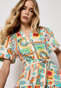 Robe courte à manches courtes colorée, avec un motif floral et géométrique vibrant, fabriquée en tissu léger avec un design croisé et une ceinture.