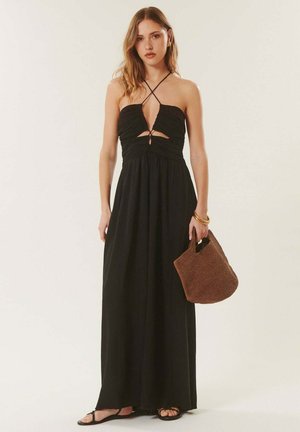 Femme portant une robe maxi noire à col halter avec découpes, tenant un sac à main tressé marron, et portant des sandales noires et des bracelets dorés.