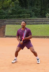 Muški tenisač u ljubičastom sportskom odijelu, drži reketa s crvenim i plavim drškom, spreman na zemljanoj podlozi okružen zelenilom.