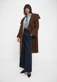 Hnědý přes oversized vlněný kabát s širokými klopami, přes světle modrou košili s knoflíky a námořnickými kalhotami s vysokým pasem. Černé boty s špičatou špičkou.