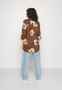 Persona con capelli ricci che indossa una blusa floreale color ruggine, jeans larghi azzurri e sneakers bianche, in piedi di spalle su uno sfondo semplice.
