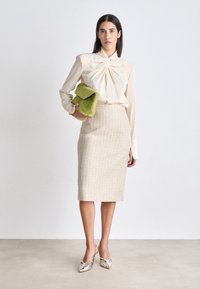 Cremefarvet bluse med tvistet front, parret med en tekstureret beige pencil skirt og sølvfarvede højhælede sko. Holder en grøn ruskindsclutch.