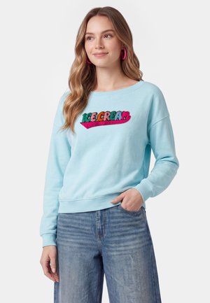 Ung kvinde med langt hår iført lyseblå sweatshirt med farverig "ICE CREAM" tekst og blå jeans, står med den ene hånd i lommen.