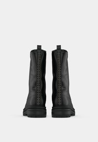 Par de botas negras de media caña con suelas gruesas, tiradores traseros visibles y filas verticales de pequeños tachuelas doradas sobre un fondo blanco liso.