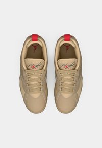 Baskets de sport beiges avec une tige synthétique lisse, des accents rouges sur le talon et la languette, et des détails en mesh. Dispose de lacets et d'un logo signature.