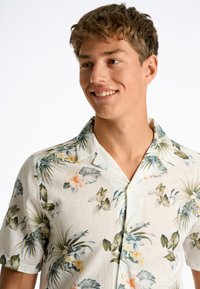 Camicia a maniche corte in tessuto leggero con una base bianca, decorata con foglie tropicali verdi e motivi floreali colorati. Collo classico e chiusura con bottoni.