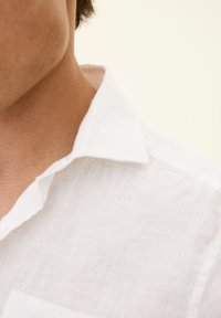 Camicia di lino bianca con una superficie texturizzata, colletto con bottoni e cuciture visibili, posizionata attorno all'area del collo e della spalla.