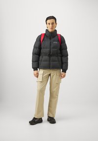 Czarna kurtka puchowa z wysokim kołnierzem i paskiem, khaki spodnie cargo z kieszeniami, czerwona plecak oraz czarne buty trekkingowe na jednolitym tle.