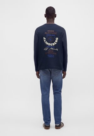 Mand stående med ryggen mod kamera iført navy langærmet skjorte med trykt tekst og grafik samt blå jeans mod en hvid baggrund.