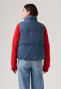 Donkerblauwe denim puffer vest met gestikt patroon, hoge kraag en knoopsluiting, gedragen over een rode ribbed trui en lichtblauwe jeans.