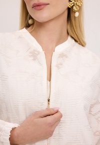 Biała bluzka o fakturze z złotym zamkiem i okrągłym dekoltem. Ręka modelki trzyma zamek, eksponując szczegóły i dopasowanie odzieży.