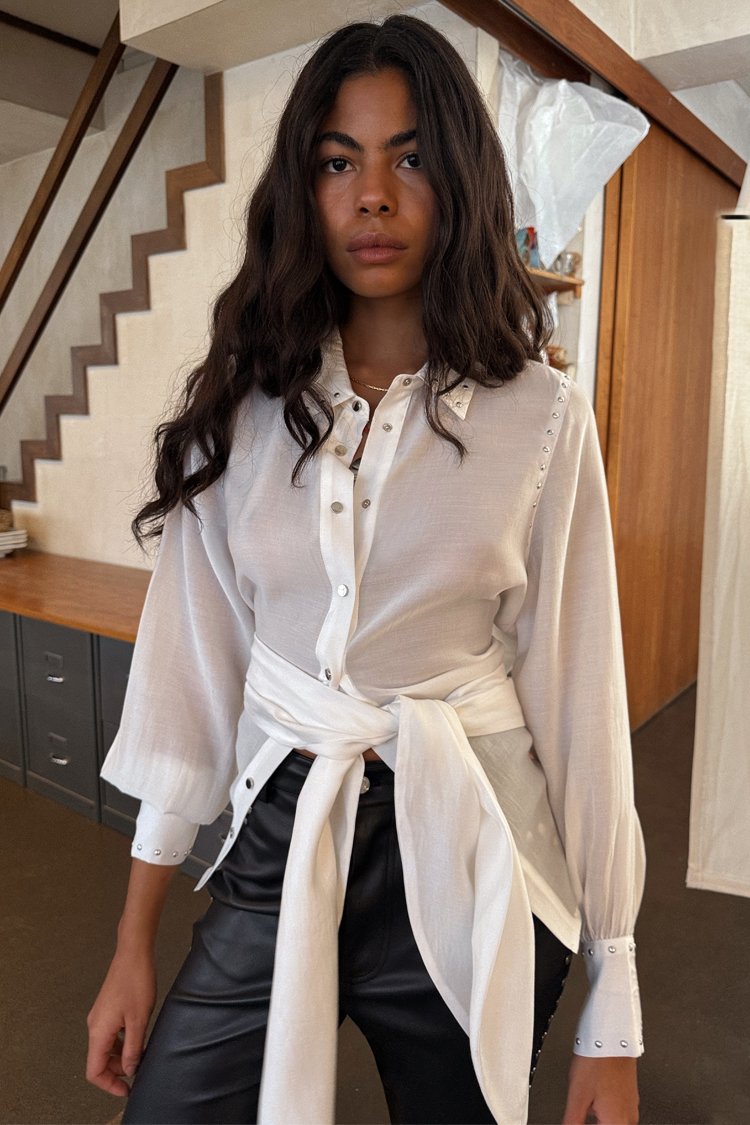 White long-sleeve blouse with a tied waist, featuring button details and textured fabric. Paired with black leather pants.