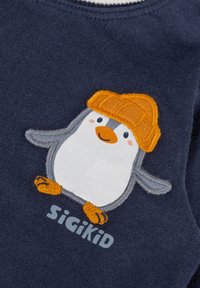 Dunkelblaues Material mit einer Pinguin-Stickerei, der einen orangefarbenen Strickhut und orangefarbene Füße trägt; unter dem Design befindet sich das "Sigikid"-Logo in Grau.