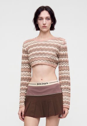 Frau mit kurzen dunklen Haaren trägt einen rosa-beigen, wellenförmig gemusterten Cropped-Pullover und einen braunen plissierten Rock mit Palm Angels Bund.