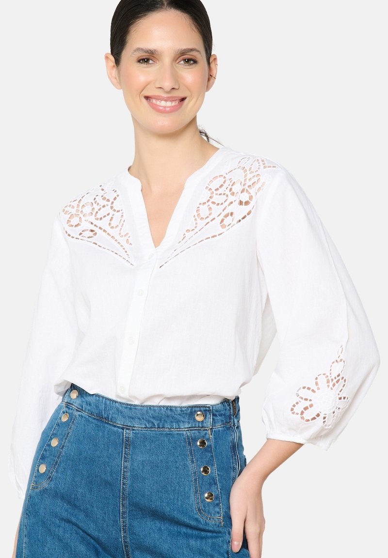 Blusa blanca de botón con bordado recortado en los hombros y las mangas, combinada con una falda de mezclilla azul de tiro alto con acentos de botones dorados.
