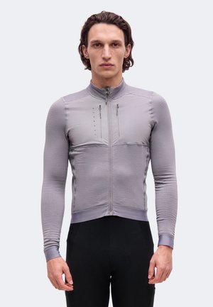 Un homme porte une veste de cyclisme grise ajustée, zippée et dotée de poches poitrine, ainsi qu’un cuissard noir, sur un fond clair et uni.
