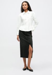 Chemise blanche à col avec un design peplum, associée à une jupe en denim noire taille haute avec une fente latérale et des chaussures plates noires.