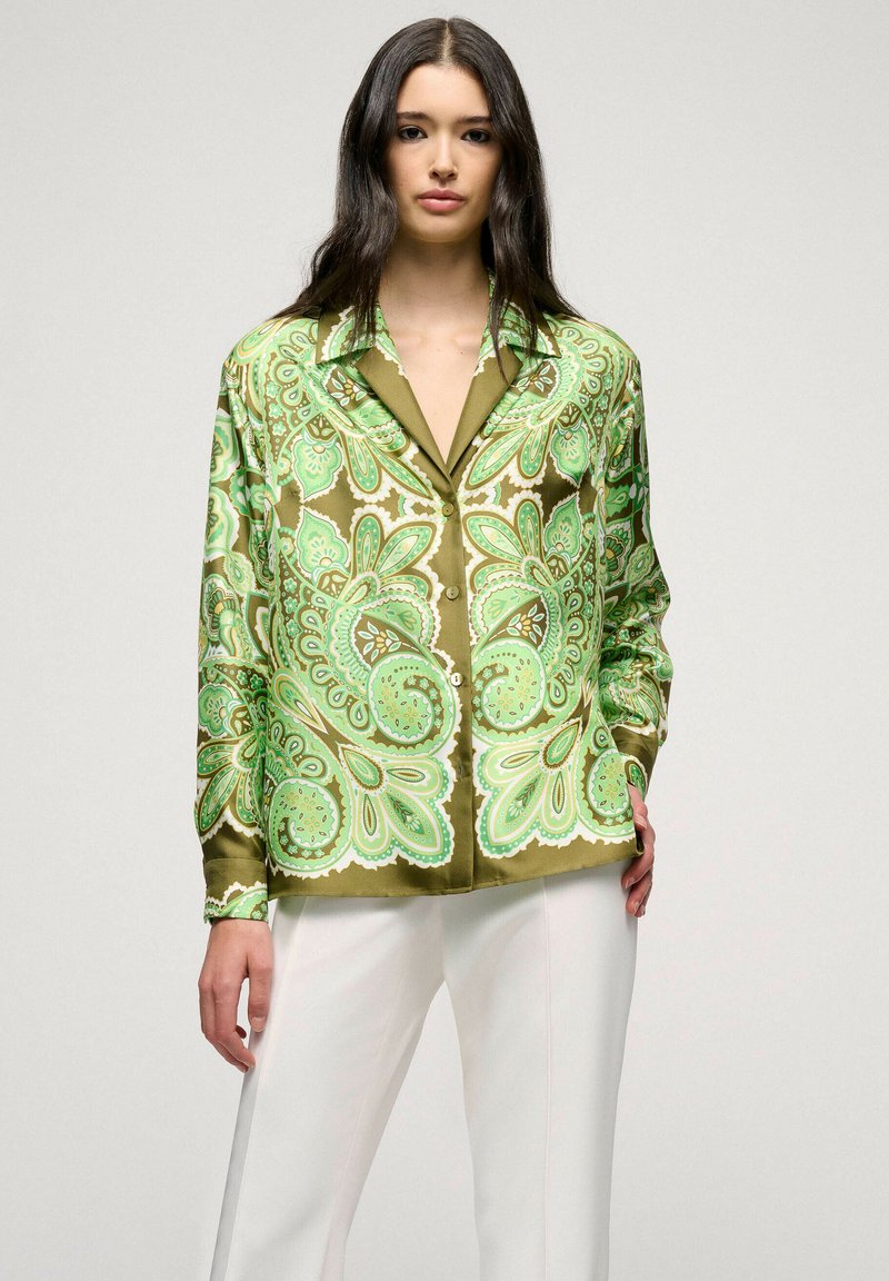 Chemise en satin vert à motif cachemire avec un col pointu, fermeture à boutons et manches longues, assortie à un pantalon blanc.