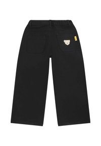 Schwarze weit geschnittene Hosen mit zwei Gesäßtaschen, wobei eine mit einem gestickten Bärenmotiv verziert ist und ein gelbes Etikett am Bund angebracht ist.