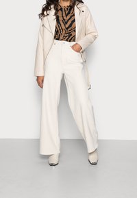 Giacca da biker in pelle sintetica crema sopra una maglietta a stampa zebra marrone, abbinata a pantaloni bianchi a gamba larga e stivaletti beige con accenti in argento.