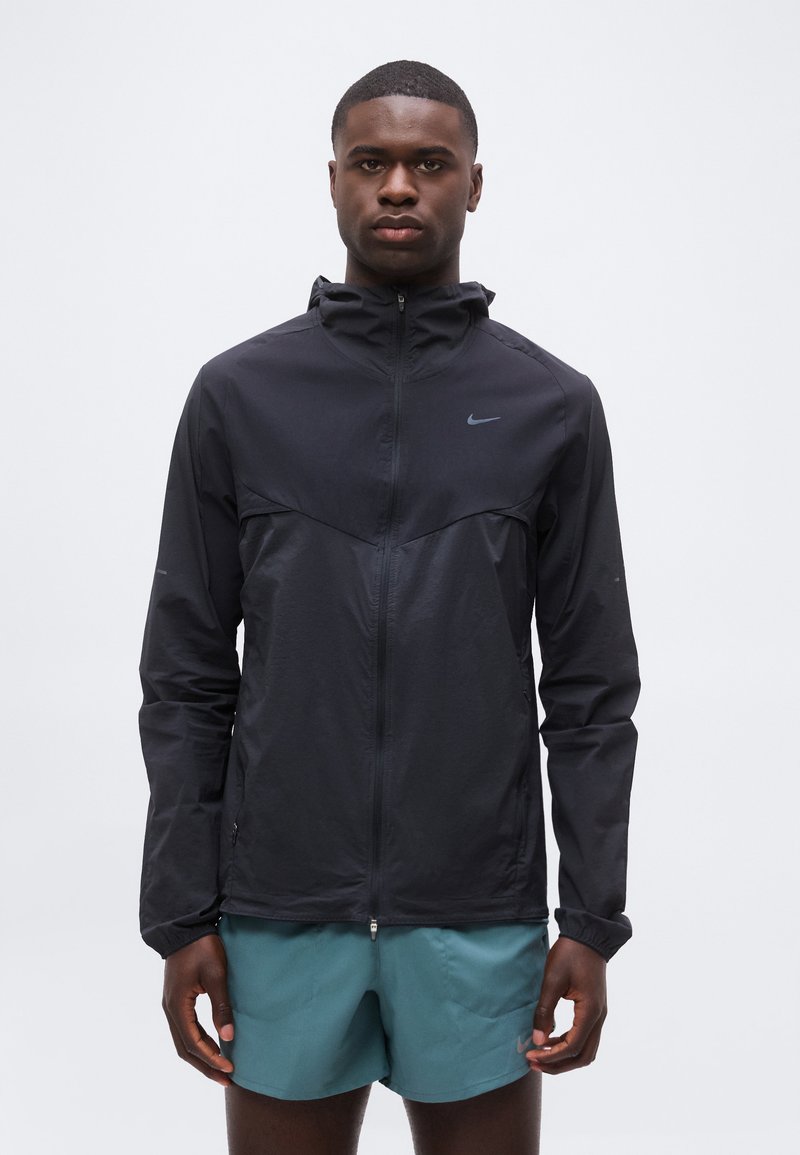 Joven con una chaqueta negra con capucha y cremallera y pantalones cortos de correr en color azul verdoso, de pie frente a un fondo claro y sencillo.