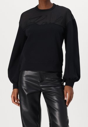 Haut noir à manches longues avec empiècement transparent et broderie "Karl", associé à un pantalon noir en simili cuir, porté par une personne debout.