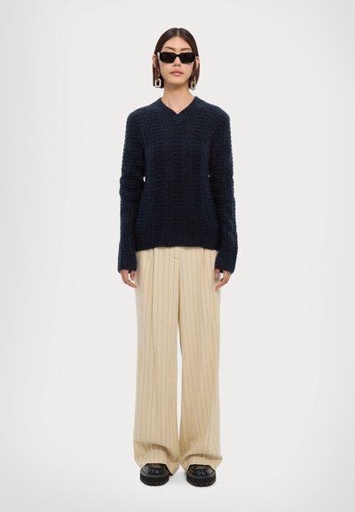 Navy knitted sweater with a V neckline, paired with beige pinstriped wide-legged trousers and black chunky shoes. Black sunglasses and earrings.