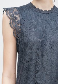 Pelēks mežģīņu tops ar ziedu rakstiem, viļņotu kakla izgriezumu un teksturētām mežģīņu piedurknēm ar pomponu akcentiem apakšmalā.