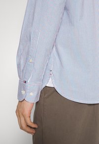 Chemise à manches longues de couleur bleu clair avec des rayures rouges et blanches, présentant des poignets boutonnés et un ourlet arrondi. Fabriquée en tissu tissé, mettant en valeur des lignes nettes.