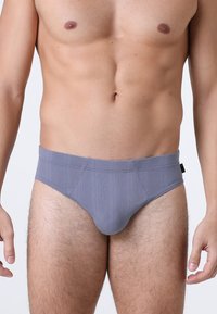 Uomo che indossa slip grigi a trama, in piedi con le braccia rilassate lungo i fianchi su uno sfondo chiaro e semplice.