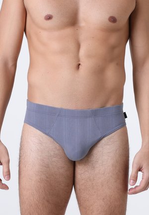 Uomo che indossa slip grigi a trama, in piedi con le braccia rilassate lungo i fianchi su uno sfondo chiaro e semplice.