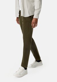 Olive green corduroy pants with a tapered fit. Paired with white sneakers and a light striped button-up shirt. Textured fabric visible.