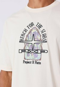 Crème katoenen t-shirt met een kleurrijk ontwerp in de stijl van een glas-in-loodraam, met de tekst "Reach for the Stars" en de logo's "PXP" en "Project X Paris."