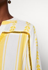 Persona vestida con una prenda de rayas blancas y amarillas, con patrones bordados y detalles calados cerca del hombro y la manga.