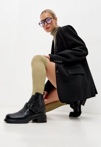 Botas negras de cuero hasta el tobillo con un detalle de hebilla, combinadas con calcetas de canalé verde oliva hasta la rodilla y una blazer negra oversized.