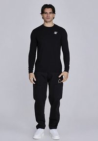 Schwarzes langärmeliges Hemd mit kleinem weißem Logo auf der Brust, kombiniert mit schwarzen Cargo-Hosen mit Seitentaschen und einem goldfarbenen Akzent.