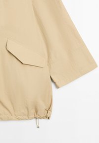 Manche de veste beige avec une poche à rabat et un ourlet à cordon de serrage réglable sur un fond blanc uni.