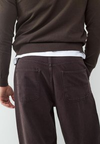 Bruine gebreide trui over een witte blouse, gecombineerd met donkerpaarse broek met twee achterzakken en een rechte pasvorm.