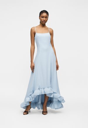 Femme portant une robe bleu clair à ourlet asymétrique à volants, avec des fines bretelles et des talons noirs ouverts, debout devant un fond blanc uni.