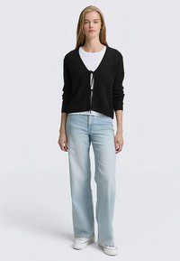 Zwarte, cropped cardigan met een striksluiting aan de voorkant, gecombineerd met een wit T-shirt en lichtblauwe uitlopende jeans. Relaxte pasvorm, zachte textuur.