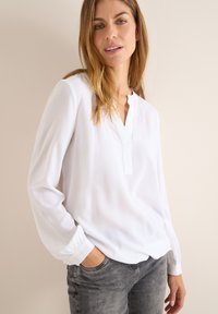 Hvid langærmet bluse med v-halsudskæring, letvægtsstof og samlede manchetter, parret med grå jeans. Glat tekstur, afslappet design.