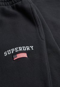 Zwarte stof met de tekst "SUPERDRY" boven een geborduurde Amerikaanse vlag nabij de rand van de zak op een paar broeken of shorts.