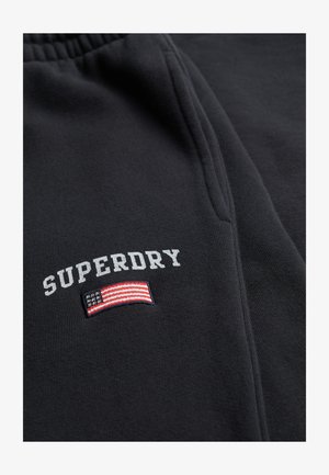 Zwarte stof met de tekst "SUPERDRY" boven een geborduurde Amerikaanse vlag nabij de rand van de zak op een paar broeken of shorts.