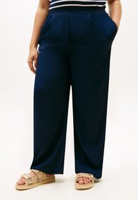 Μπλε-σκούρο παντελόνι με φαρδιά μπατζάκια, πλισέ μπροστινό μέρος, πλαϊνές τσέπες και λεία υφή. Φορεμένο με ταμπά σανδάλια που διαθέτουν διακοσμητικό σχεδιασμό λουριού.