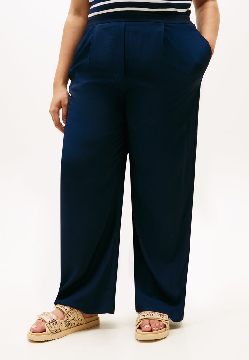 Μπλε-σκούρο παντελόνι με φαρδιά μπατζάκια, πλισέ μπροστινό μέρος, πλαϊνές τσέπες και λεία υφή. Φορεμένο με ταμπά σανδάλια που διαθέτουν διακοσμητικό σχεδιασμό λουριού.