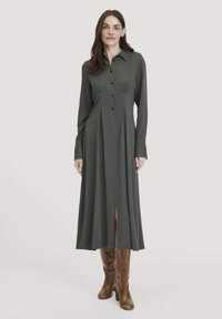 Frau in einem langärmligen, dunkelgrauen, knöpfbaren Kleid mit Frontfalten und braunen knielangen Stiefeln, die vor einem schlichten Hintergrund steht.