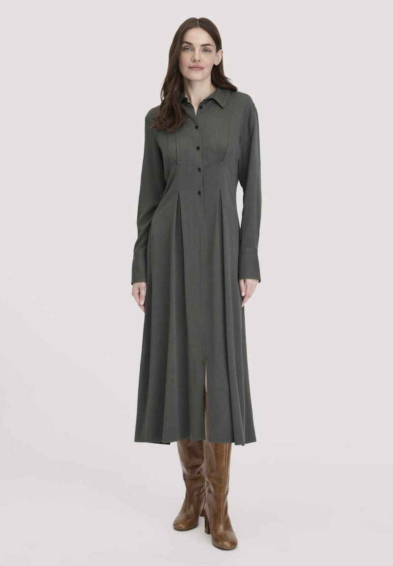 Frau in einem langärmligen, dunkelgrauen, knöpfbaren Kleid mit Frontfalten und braunen knielangen Stiefeln, die vor einem schlichten Hintergrund steht.