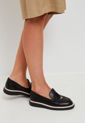 Frau trägt schwarze Leder-Loafer mit silberner Schnallenverzierung, kombiniert mit einem beigen Rock, steht auf einem weißen Boden.